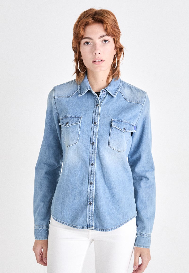 Camisa de mezclilla azul claro con cuello, mangas largas y dos bolsillos en el pecho. Presenta cierre de botones y un dobladillo curvado.
