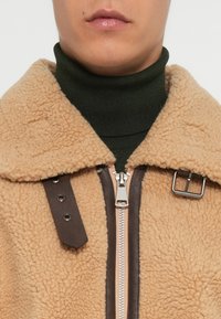 Beige shearling jakke med en brun læderrem detalje, sølv lynlås og en grøn turtleneck under. Blød tekstur og kravedesign.