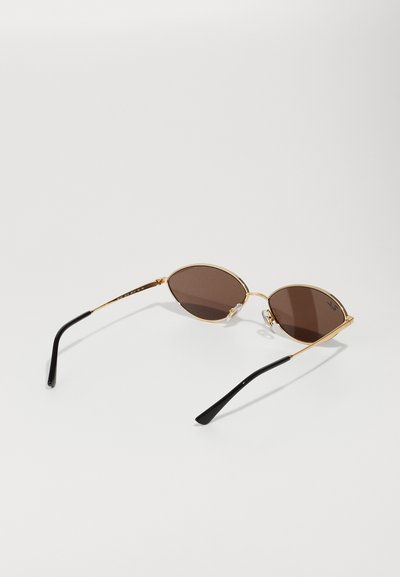 Des lunettes de soleil ovales avec une monture dorée, des verres marron, des coussinets de nez transparents, et des embouts de branches noirs, présentant un design mince et peu d'ornementation.
