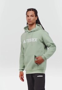Man med flätat hår som bär en ljusgrön Adidas Terrex-hoodie och svarta byxor, med en hand i framfickan mot en enkel bakgrund.