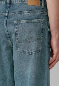 Jasnoniebieskie dżinsy z tylną kieszenią z subtelnym detalem w formie falistych szwów oraz z brązową skórzaną metką powyżej, prezentującą teksturowane wykończenie.