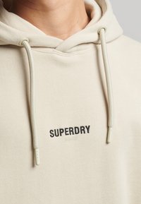 Mikina s kapucí ve světlém béžovém textilu, s černým vyšitým logem "SUPERDRY" a texturovanými šňůrkami se plastovými konci.