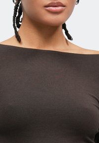 Καφέ τοπ με ανοιχτούς ώμους από απαλό, ελαστικό ύφασμα με ομαλή υφή. Η κοντινή λήψη αναδεικνύει το λαιμό και το σχέδιο στους ώμους.