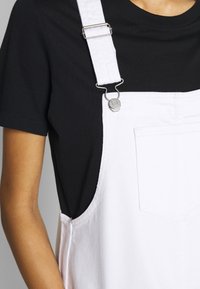 Bílé denimové overaly s nastavitelnými ramínky, stříbrnými detaily a velkou přední kapsou, v kombinaci s černým tričkem. Hladká textura, ležérní design.