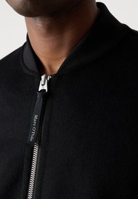 Black bomber jacket with a smooth fleece texture, ribbed collar, and a silver zipper. Features a white logo tag on the zipper pull.