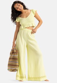 Light yellow two-piece outfit featuring a smocked crop top with a ruffled hem and wide-leg pants with lace trim. Model holds a multicolored bag.