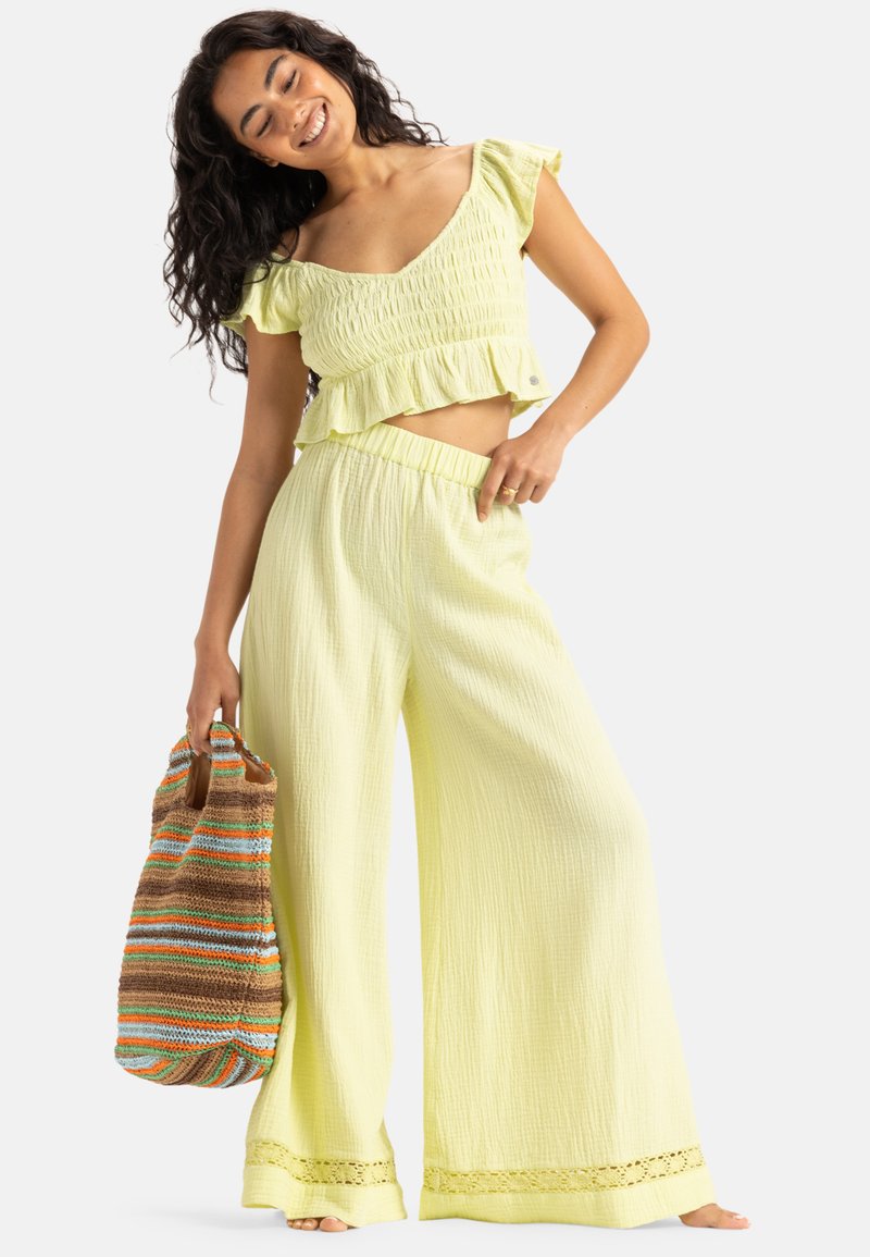 Light yellow two-piece outfit featuring a smocked crop top with a ruffled hem and wide-leg pants with lace trim. Model holds a multicolored bag.