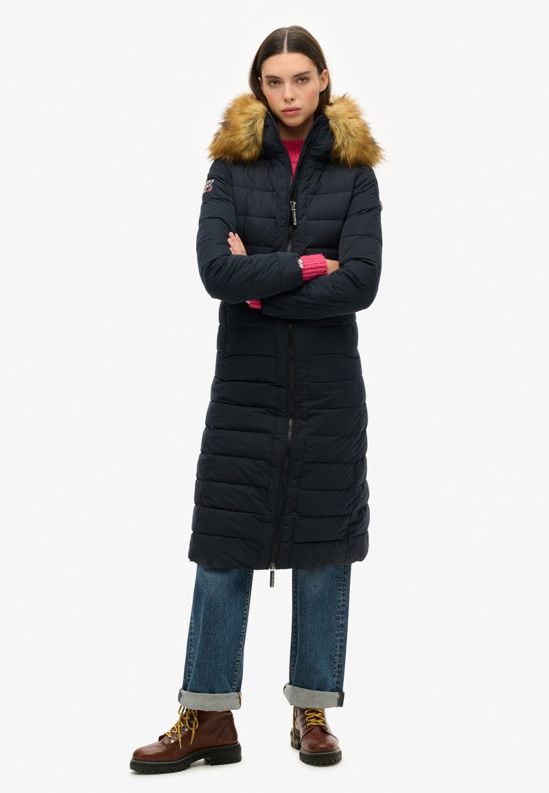 Marineblauwe donsjas met bontomzoomde capuchon, ritsluiting, lange lengte en horizontaal gestikte design. Gecombineerd met een roze gebreide trui en denim.