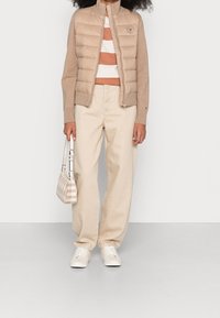 Lichtbeige puffer vest met een hoge kraag, gecombineerd met een gestreepte top in bruin en wit, lichtbeige broek en een kleine handtas met ruitpatroon.