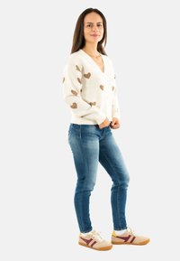 Donna in piedi che indossa un cardigan bianco con cuori marroni, jeans blu e sneakers beige con strisce bordeaux su uno sfondo bianco.
