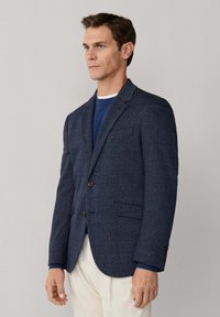 Muž v námořnicky modrém kostkovaném saku přes modrý svetr a bílé kalhoty, stojící před jednoduchým světle šedým pozadím.