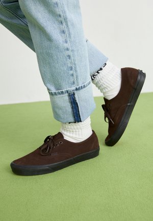 Bruine suède sneakers met zwarte rubberen zolen, veters en veterogen, gecombineerd met witte geribbelde sokken en lichtblauwe opgerolde jeans.