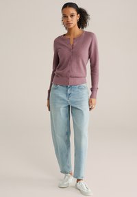 Lichtpaarse cardigan met lange mouwen en textuurpatroon, met knopen aan de voorkant, gecombineerd met lichtblauwe hoogwater jeans en witte schoenen.