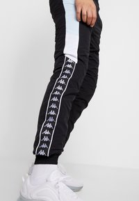 Svarta träningsbyxor med sidopaneler i ljusblått och vitt, med ett vertikalt logomönster ner längs yttersemen. Elastiska muddar och midja.