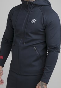 Mörkblå zip-up huvtröja i slät tyg, med en framsideslogotyp, sidofickor med dragkedja och röd detalj på ärmen. Justerbar dragsko för huvan.