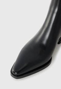 Bottine en cuir noir avec un bout pointu, texture lisse et un talon fin. La semelle présente un minimum de détails pour un aspect élégant.