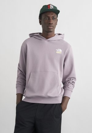 Man draagt een lichtpaarse The North Face-hoodie en donkere broek, met handen in de zakken en een groene baseballpet naar voren gericht.