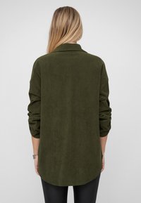 Donkergroen corduroy overhemd met een kraag, lange mouwen en een afgeronde zoom. De stof heeft een geribbelde textuur.