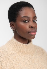 Ung sort kvinde med kort krøllet hår iført en beige strikket sweater, små hoop- og studøreringe, poserer mod en hvid baggrund.