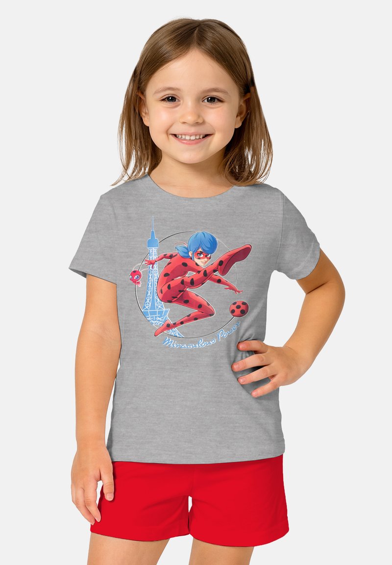 Young girl smiling, wearing gray t-shirt with animated superhero in red and black spots, blue hair, Eiffel Tower, and red shorts.