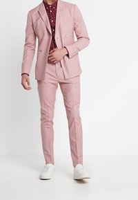 Completo rosa chiaro con giacca doppiopetto e pantaloni slim-fit, abbinato a una camicia bordeaux a fantasia e scarpe da ginnastica bianche.