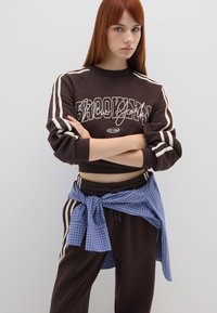 Sudadera corta marrón con gráfico de "BROOKLYN" y franjas crema en las mangas. Joggers marrones que presentan franjas laterales y una camisa de cuadros azul atada en la cintura.