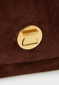Sac à main en suède marron avec un fermoir rond en métal doré et un logo gravé. La texture est douce, avec des coutures soignées le long des bords.