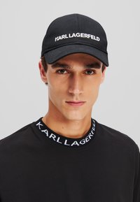 Schwarze Baumwollkappe mit gebogenem Schild, versehen mit "KARL LAGERFELD" in weißer Bestickung. Getragen mit einem passenden schwarzen Hemd und Logo-Kragen.
