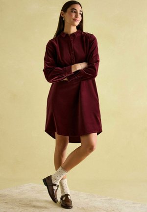 Burgundy button-up dress made of soft fabric, featuring long sleeves, gathered shoulder details, and a curved hem. Worn with dotted socks and brown shoes.