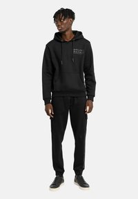 Zwarte hoodie met logo, trekkoord en voorzak; gecombineerd met zwarte joggers met cargozakken en zijlogo; zwarte sneakers.