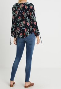 Blommig blus med långa ärmar i marinblått med rosa och gröna mönster, med en nyckelhålsspecifikation bak och bandknutar vid ärmarna. Matchad med blå jeans.
