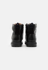 Un paio di stivali alla caviglia neri in pelle con linguette e chiusure con cerniera, visti dal retro su uno sfondo bianco.