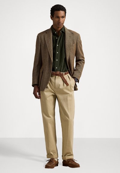 Blazer en tweed marron, chemise verte à boutons, pantalon beige, ceinture tressée marron et chaussures marron. Design formel avec des tissus texturés et des couleurs neutres.