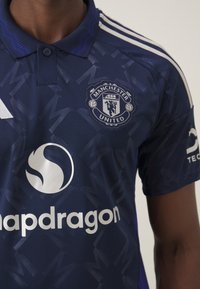 Ciemnoniebieski żer Manchesteru United z teksturowanym wzorem, haftowanym logo i białym brandingiem sponsorów. Posiada trzy białe paski na ramionach.