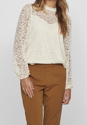 Blusa de encaje en marfil con mangas largas transparentes, patrón floral texturizado, cuello alto y puños elásticos, combinada con pantalones marrones.