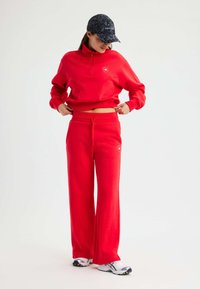 Roter Trainingsanzug mit einem Reißverschluss und hohem Kragen sowie lockeren Hosen. Der Pullover hat ein kleines Logo; die Hose verfügt über einen Kordelzug am Bund.