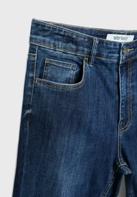 Dunkelblaue Jeans aus Denim mit glatter Textur, ausgestattet mit einer Fronttasche und kontrastierendem gelben Nähten. Markenlabel am Bund sichtbar.
