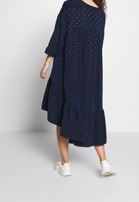 Marinblå klänning med prickar, trekvartslånga ärmar, flerlagad volangkant, mjukt tyg och en avslappnad siluett. Bärs med vita sneakers.