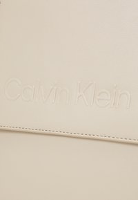 Sac en cuir beige Calvin Klein avec logo embossé, texture lisse, lignes épurées et détails de couture subtils.