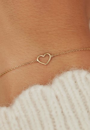 Goldene Kettenarmband mit einem hohlen Herzanhänger, vor einem strukturierten Hauthintergrund mit einem hellen Strickpullover im Blick.