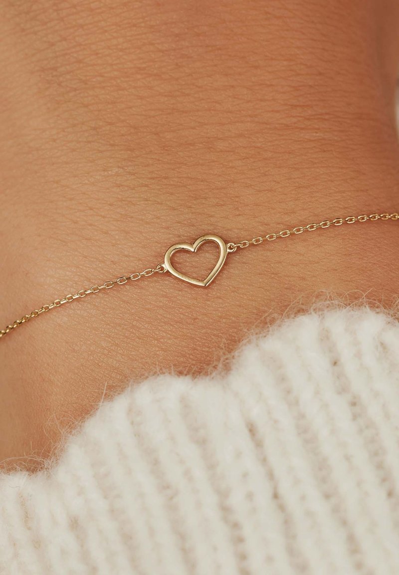Gouden kettingarmband met een holle hartbedel, tegen een achtergrond van textuur van de huid met een lichtgekleurde gebreide trui in beeld.