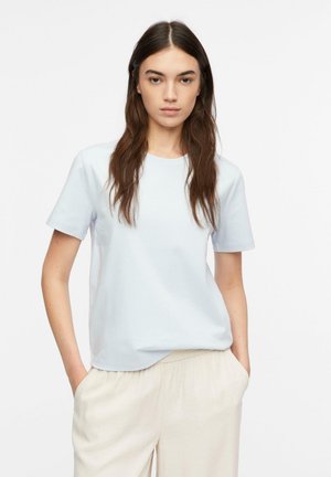 Kobieta z długimi brązowymi włosami, ubrana w jednolitą białą koszulkę z krótkim rękawem i jasnobeżowe spodnie, stojąca z rękami w kieszeniach na białym tle.