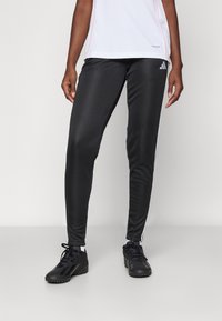 Black athletic pants with a smooth texture, featuring white side stripes, paired with black sports shoes. The shirt is white with pink accents.