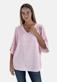 Lichtroze top met een V-hals en brede mouwen, gemaakt van zachte stof. Heeft een losse pasvorm en een gladde textuur, gecombineerd met een blauwe spijkerbroek.