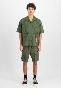 Cămașă verde cu mânecă scurtă, dotată cu două buzunare la piept și un etichetă, asortată cu pantaloni scurți cargo de culoare măslinie, care au multiple buzunare și un cordon ajustabil.
