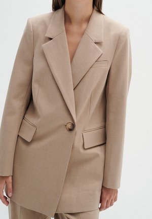 Femme portant un blazer beige à un bouton avec des revers crantés et des poches à rabat, debout devant un fond blanc uni.