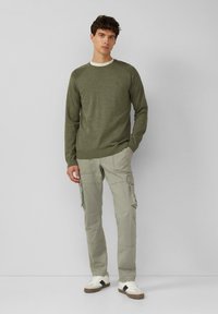 Zielony sweter z dzianiny, z ryflem na mankietach, w połączeniu z jasnoszarymi spodniami cargo z wieloma kieszeniami i luźnym krojem. Casulowe białe sneakersy.