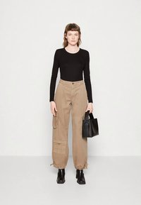 Body de mangas compridas preto combinado com calças cargo bege com bolsos cargo e bainhas com cordão. Modelo segura uma pequena mala preta.