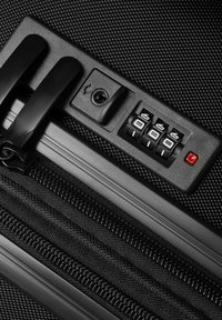 Matériel de valise noire avec un cadenas à combinaison, une glissière, et une surface texturée, avec des poignées lisses et un voyant lumineux rouge.