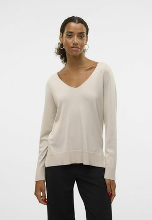 Maglione beige a V con maniche lunghe, orlo a coste e spacchi laterali; tessuto morbido e vestibilità comoda, abbinato a pantaloni neri.